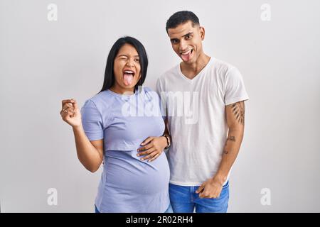 Young hispanic couple expecting a baby standing over background sticking tongue out happy with funny expression. emotion concept. Stock Photo