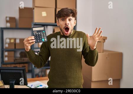 Arab man with beard working at small business ecommerce holding calculator celebrating victory with happy smile and winner expression with raised hand Stock Photo