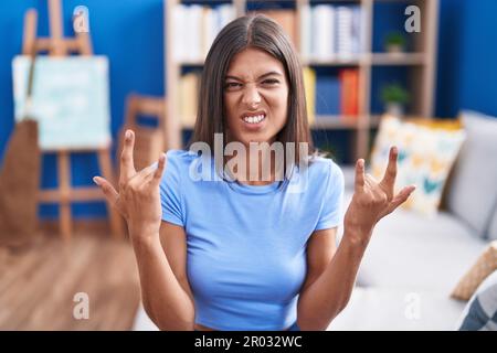 Brunette young woman sitting on the sofa at home shouting with crazy expression doing rock symbol with hands up. music star. heavy concept. Stock Photo