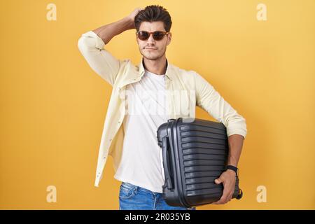 Young hispanic man holding suitcase going on summer vacation confuse and wondering about question. uncertain with doubt, thinking with hand on head. p Stock Photo