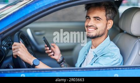 Young hispanic man using smartphone sitting on car at street Stock Photo