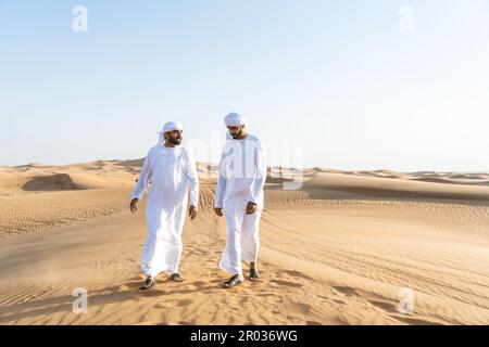 Two middle-eastern men wearing traditional emirati arab kandura bonding in the desert - Arabian muslim friends meeting at the sand dunes in Dubai Stock Photo