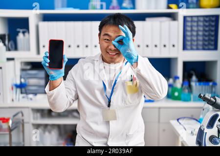 Young chinese man working at scientist laboratory holding smartphone smiling happy doing ok sign with hand on eye looking through fingers Stock Photo