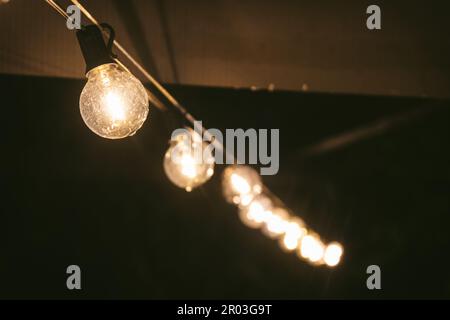 From below soft focus on row of illuminated light bulbs hanging on rope under ceiling in dark room Stock Photo