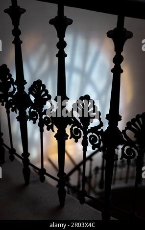 Decorative black wrought iron banister detail. Selective focus. Spiral staircase. Colored window glass reflection on the wall in the background. Stock Photo