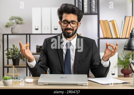 Indian businessman working on laptop computer, meditating, doing yoga breathing exercise in lotus position at home office. Calm serene freelancer arab man taking break. Busy occupation. Peace of mind Stock Photo