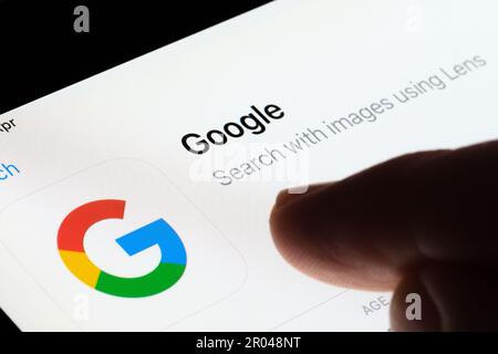 Google search app seen in App Store on the screen of ipad and blurred finger pointing at it. Selective focus. Stafford, United Kingdom, May 6, 2023 Stock Photo