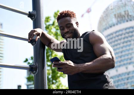 Happy fit sporty young black man using phone standing in workout park. Stock Photo