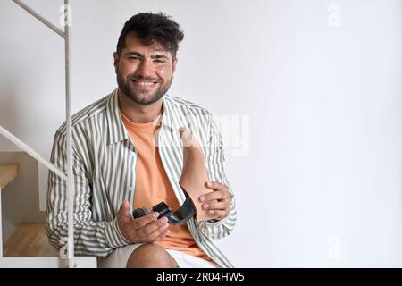 Young happy man with disability holding foot leg prosthesis sitting on stairs. Stock Photo