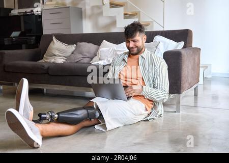 Happy man with disability leg prosthesis sitting on floor at home using laptop. Stock Photo