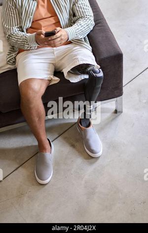Amputee man with leg prosthesis sitting on sofa at home using phone. Stock Photo