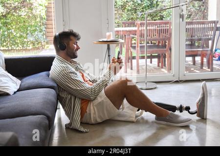 Happy man with leg prosthesis at home wearing headphones using phone. Stock Photo