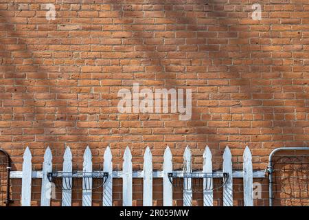 Abstract texture background of a vintage red exterior brick wall with weathered bricks and diagonal line shadows in sunlight, and a rustic wood fence Stock Photo