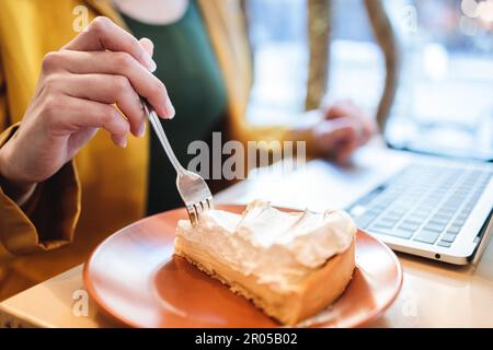 Unrecognizable remote worker enjoying lemon pie while using laptop in a yellow blazer at a cafe. Digital Nomad. Stock Photo