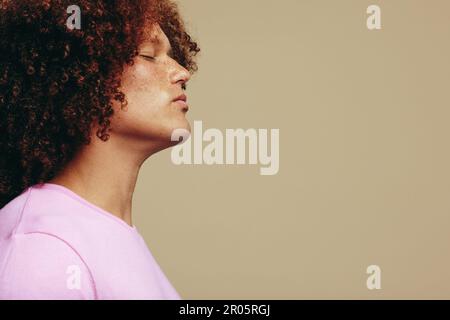 Young man standing in a studio with his eyes closed, embracing his freckled skin and ginger afro hair. Youthful and handsome man expressing confidence Stock Photo