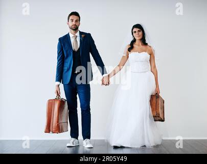 We have our travel shoes on. Studio shot of a newly married couple holding hand and carrying bags while standing against a gray background. Stock Photo