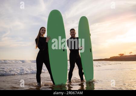 Smiling Sporty Couple In Wetsuits Posing With Surfboards On Beach At Sunset Stock Photo