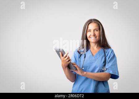 Friendly senior woman doctor in uniform posing with stethocsope and holding digital tablet, using medical mobile app Stock Photo