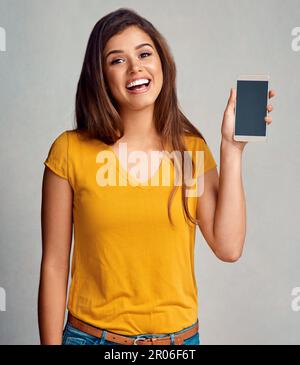 Hey Check this out. Studio portrait of an attractive young woman holding a cellphone with a blank screen against a grey background. Stock Photo