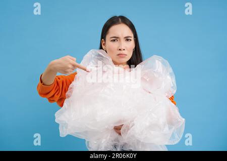Angry eco-activist points at big pile of plastic bags, fighting to save environment, encourage to recycle and go green, blue background Stock Photo