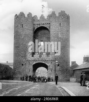 A late 19th century view of the Saint Laurence Gate, built in the 13th century as part of the walled fortifications of the medieval town of Drogheda in County Louth, Ireland. It is a barbican or defended fore-work which stood directly outside the original gate of which no surface trace survives. The structure consists of two towers, each with four floors, joined by a bridge at the top,  an entrance arch at street level under which there is a slot from where a portcullis originally could be raised and lowered.