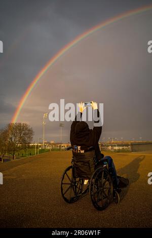 A woman wearing a black hoodie and sat in a wheelchair takes a photograph of a rainbow using her mobile phone. Location is the Olympic Park, Stratford Stock Photo