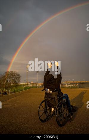 A woman wearing a black hoodie and sat in a wheelchair takes a photograph of a rainbow using her mobile phone. Location is the Olympic Park, Stratford Stock Photo