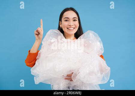 Asian woman with plastic bags, recycling waste in hands, points up, showing recycle sorting center, sustainable life tips, blue background Stock Photo