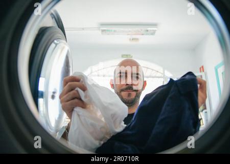 A man loads the washing machine with dirty clothes in the laundromat. Stock Photo