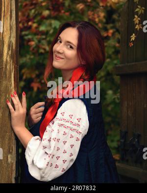 Smiling cheerful young redhead woman dressed traditional ukrainian embroidered clothes and with a red ribbon in her hair looking at the camera. Stock Photo
