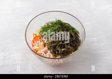 Glass bowl with shredded cabbage, grated carrots, chopped sweet peppers, red onion, seaweed and dill on light gray background. Step of preparing healt Stock Photo