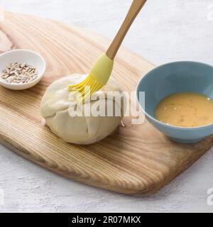 Small raw pie smeared with egg before baking surrounded by two bowls with a beaten egg and seeds on light gray background. Stage of cooking delicious Stock Photo