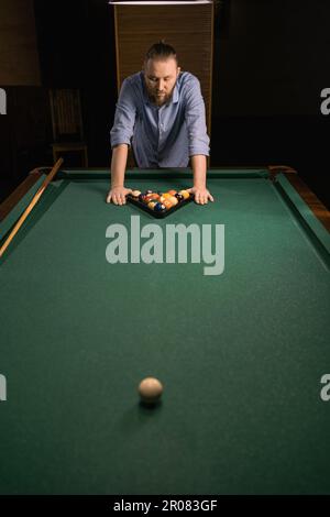 Sports game of billiards on a green cloth. Millennial man putting multi-colored billiard balls in the form of a triangle with numbers, a triangle on a Stock Photo