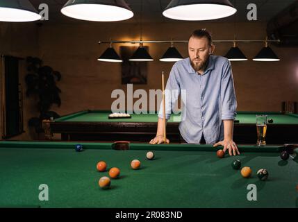 Thoughtful caucasian bearded man plays billiards, thinking and looking on pool. Billiard room on the background. Hobby, leisure and playing billiards Stock Photo