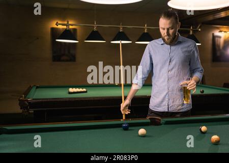 Young man playing snooker, concentrating with cue in hand, watching holding glass of beer Stock Photo