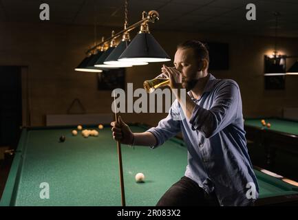 Young professional man playing billiards and drinking beer in the dark billiard club Stock Photo