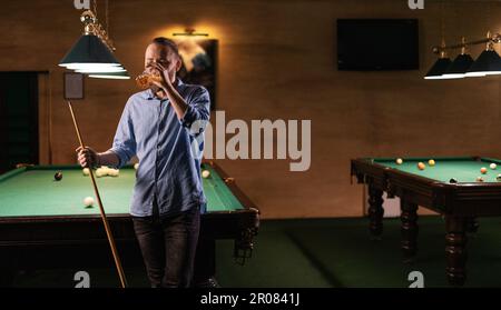 Young handsome man drinking beer beside billiard table. Male evening in billiard club. Men drinking beer and playing snooker Stock Photo