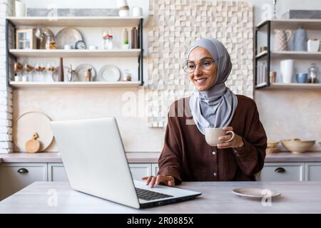 Muslim young woman in hijab sitting at the table in the kitchen at home in the morning. Drinks coffee, uses laptop, checks mail, reads news, smiles. Stock Photo