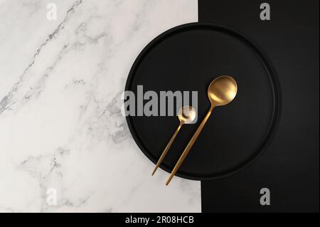 Top view of black plate and gold cutlery on white marble background. Creative table setting flat lay. Plate, spoon and dessert spoon. Copy space. Stock Photo
