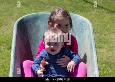 Image of two adorable smiling children holding each other inside a wheelbarrow. Brotherly love as they play in the garden Stock Photo