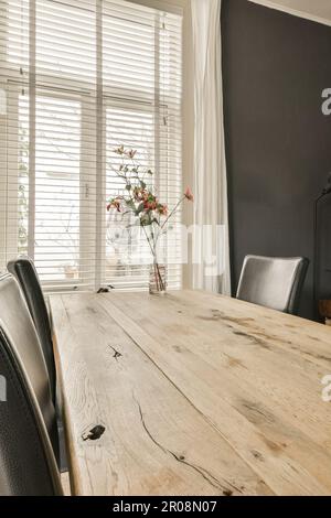 a dining table and chairs in a room with white blinds on the window behind it is a vase of flowers Stock Photo
