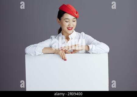 smiling elegant air hostess asian woman in red skirt and hat uniform with blank billboard isolated on grey. Stock Photo
