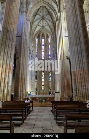 Batalha, Portugal - August 15, 2022: Interior of gothic Monastery showing altar with cross, stained-glass windows, pews and tourists Stock Photo