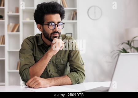 Bored millennial generation man sitting ay the table, feeling lonely at home. Unhappy young male suffering from negative thoughts, having depressive Stock Photo