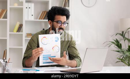 A young Indian male businessman is sitting at the table, talking on a video call on a laptop with colleagues, showing graphs, discussing work points. Stock Photo