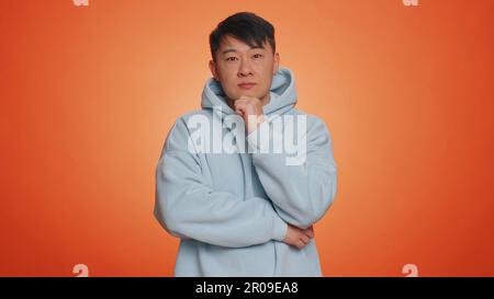 Thoughtful clever asian handsome man rubbing his chin and looking aside with pensive expression, pondering a solution, doubting question. Adult guy on orange studio background. People lifestyles Stock Photo