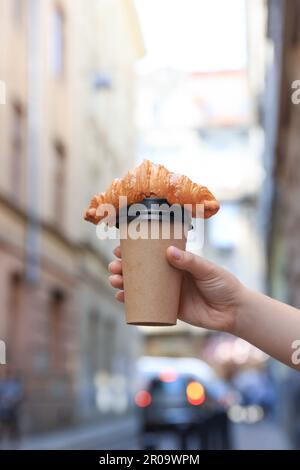 Woman holding tasty croissant and cup of coffee on city street, closeup Stock Photo