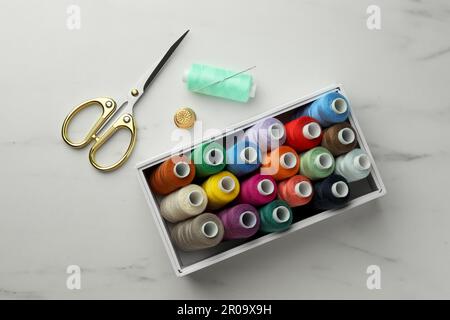 Box with colorful sewing threads, scissors and thimble on white marble table, flat lay Stock Photo