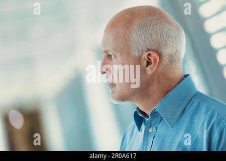 Profile view of old man in office attire looking between illuminated slits in a modern corridor of a hospital or business. He looks out and seems to b Stock Photo