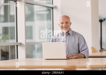 Aged man with a laptop computer in a large bright room looks at you mouthing off and making funny faces laughing, because he's young at heart and alwa Stock Photo
